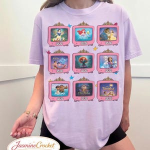 Op de afbeelding: Lichtpaars T-shirt met een raster van roze televisies, elk met een sc&egrave;ne uit een animatiefilm. De top heeft een ronde hals en korte mouwen. De tekst "Jasmine Crocket" staat onderaan.