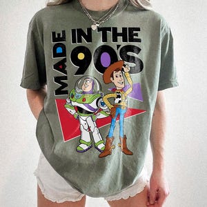 May include: An olive green t-shirt with the text "MADE IN THE 90'S" and cartoon characters Buzz Lightyear and Woody. The design includes red, purple, and white elements. The shirt has a crew neck and short sleeves.
