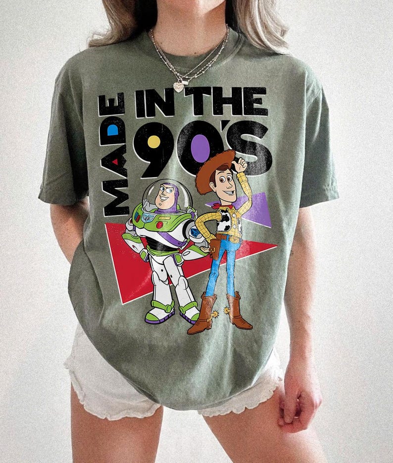 May include: An olive green t-shirt with the text "MADE IN THE 90'S" and cartoon characters Buzz Lightyear and Woody. The design includes red, purple, and white elements. The shirt has a crew neck and short sleeves.