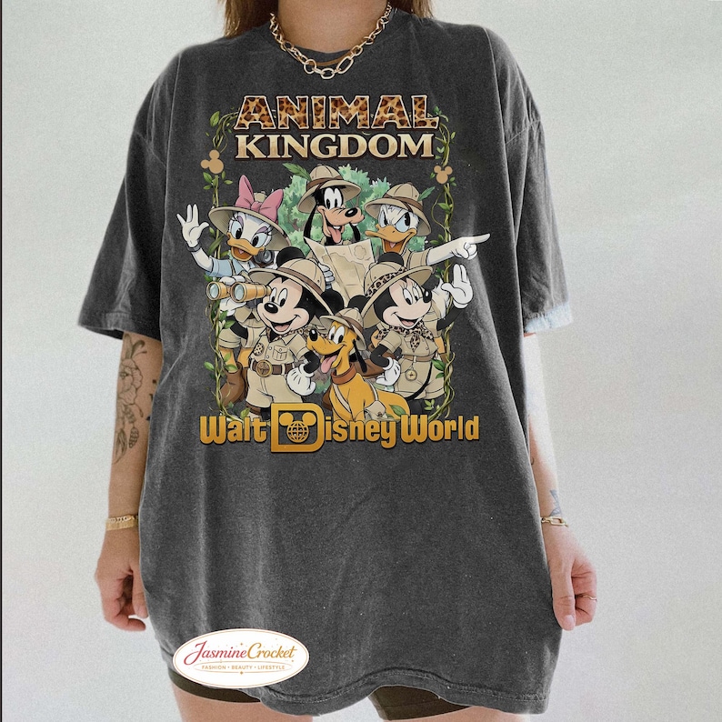 Op de afbeelding: Donkergrijze t-shirt met een afbeelding van Disney-personages in safari-outfits, waaronder Mickey Mouse, Donald Duck en Goofy. De teksten "ANIMAL KINGDOM" en "Walt Disney World" staan boven en onder de personages.