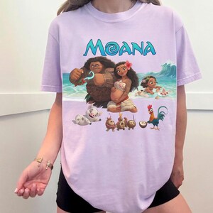 Puede incluir: Camiseta lila claro con la palabra "MOANA" en turquesa y un gr&aacute;fico de personajes de la pel&iacute;cula animada. El gr&aacute;fico incluye un hombre, una mujer, un beb&eacute;, un cerdo y un gallo en una escena de playa.