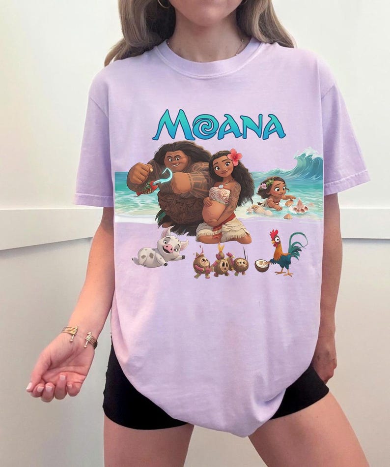 Puede incluir: Camiseta lila claro con la palabra "MOANA" en turquesa y un gr&aacute;fico de personajes de la pel&iacute;cula animada. El gr&aacute;fico incluye un hombre, una mujer, un beb&eacute;, un cerdo y un gallo en una escena de playa.