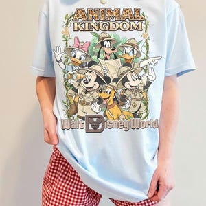 Op de afbeelding: Lichtblauw T-shirt met een afbeelding van Disney-personages in safari-outfits, met de tekst "ANIMAL KINGDOM" en "Walt Disney World". Het ontwerp bevat Mickey Mouse, Minnie Mouse, Donald Duck, Daisy Duck, Goofy en Pluto.