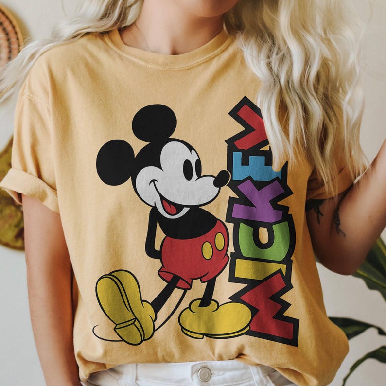 May include: Mustard yellow t-shirt with a smiling Mickey Mouse graphic. Mickey is in his classic outfit: red shorts, yellow shoes, and white gloves. The word "Mickey" is printed in colourful, stacked letters next to the character.