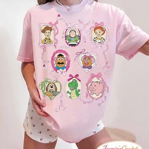 以下が含まれることがあります： ピンク色のTシャツ。フレームに入った漫画のキャラクターの肖像画が特徴で、ピンクのリボンとリボンで飾られています。キャラクターには、カウボーイ、宇宙レンジャー、カウガール、ポテトヘッド、ポテトヘッドの妻、犬、恐竜、豚が含まれます。