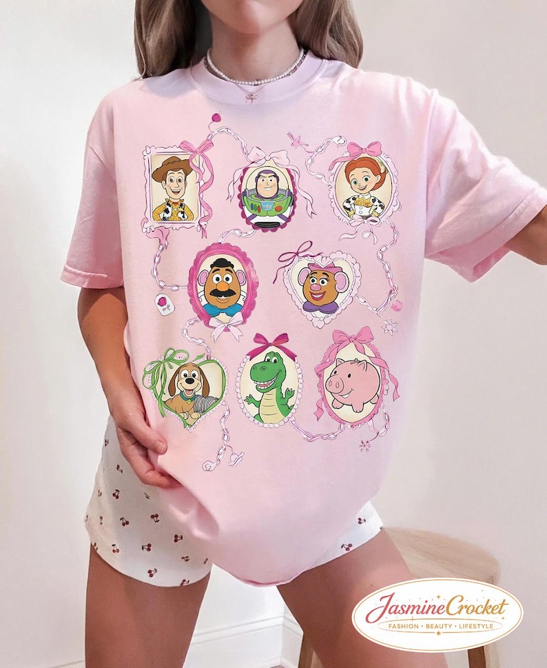 以下が含まれることがあります： ピンク色のTシャツ。フレームに入った漫画のキャラクターの肖像画が特徴で、ピンクのリボンとリボンで飾られています。キャラクターには、カウボーイ、宇宙レンジャー、カウガール、ポテトヘッド、ポテトヘッドの妻、犬、恐竜、豚が含まれます。