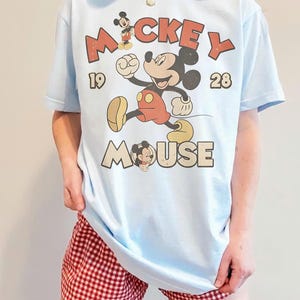 Op de afbeelding: Lichtblauw T-shirt met een vintage Mickey Mouse-afbeelding. Het ontwerp toont Mickey Mouse in beweging, de woorden "Mickey Mouse" en de cijfers "1928". Het shirt wordt gecombineerd met een rood-witte geruite short.