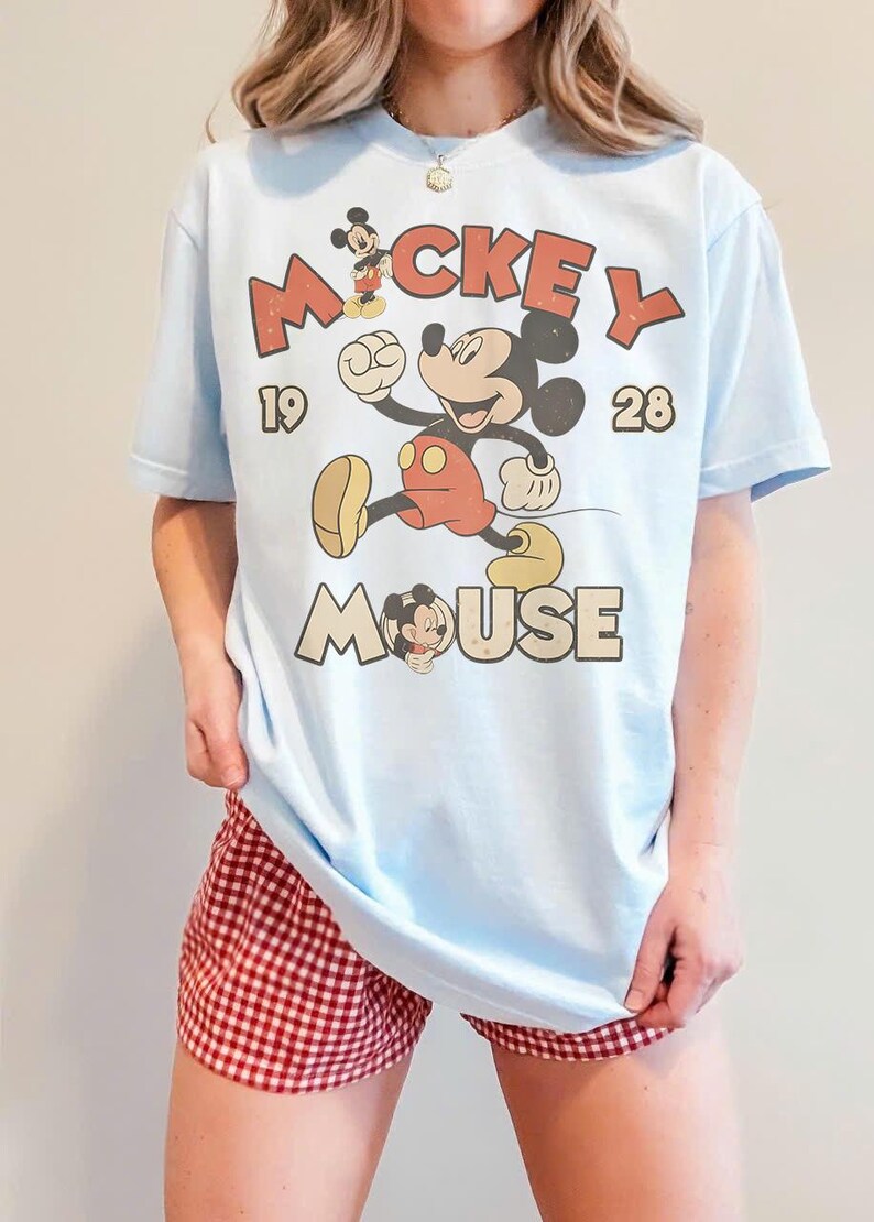 Op de afbeelding: Lichtblauw T-shirt met een vintage Mickey Mouse-afbeelding. Het ontwerp toont Mickey Mouse in beweging, de woorden "Mickey Mouse" en de cijfers "1928". Het shirt wordt gecombineerd met een rood-witte geruite short.