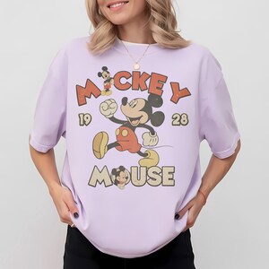 Op de afbeelding: Lichtpaars T-shirt met een vintage Mickey Mouse-ontwerp. De afbeelding toont Mickey Mouse die rent, met de woorden "Mickey Mouse" en de cijfers "1928". Het shirt heeft korte mouwen.