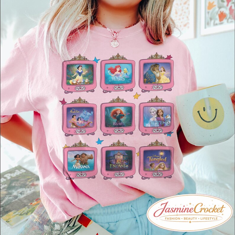 Op de afbeelding: Roze T-shirt met een raster van negen roze televisies, elk met een andere animatiefilmsc&egrave;ne. Het shirt heeft een ronde hals en korte mouwen. Een witte mok met een gele smiley wordt in de rechterhand gehouden.