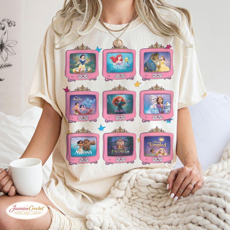 Op de afbeelding: Cr&egrave;mekleurig T-shirt met roze televisieschermen met afbeeldingen van geanimeerde prinsessen. De schermen tonen personages uit populaire animatiefilms. Het shirt heeft een relaxte pasvorm en korte mouwen.