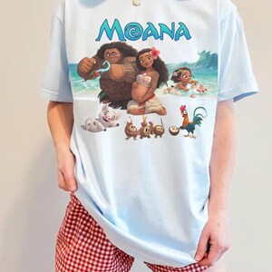 Puede incluir: Camiseta azul claro con la palabra "MOANA" en escritura azul y un gr&aacute;fico colorido de personajes de la pel&iacute;cula animada. La camiseta se usa con pantalones cortos a cuadros rojos y blancos.