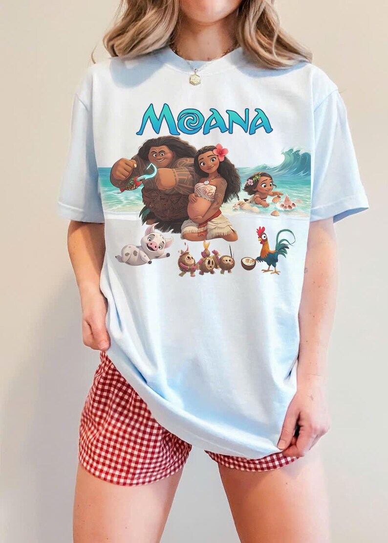 Puede incluir: Camiseta azul claro con la palabra "MOANA" en escritura azul y un gr&aacute;fico colorido de personajes de la pel&iacute;cula animada. La camiseta se usa con pantalones cortos a cuadros rojos y blancos.