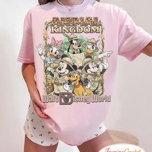 Op de afbeelding: Lichtroze T-shirt met een afbeelding van Disney-figuren in safari-outfits. De tekst "ANIMAL KINGDOM" staat bovenaan, met "Walt Disney World" eronder. Het ontwerp bevat Mickey Mouse, Minnie Mouse, Donald Duck, Daisy Duck, Goofy en Pluto.