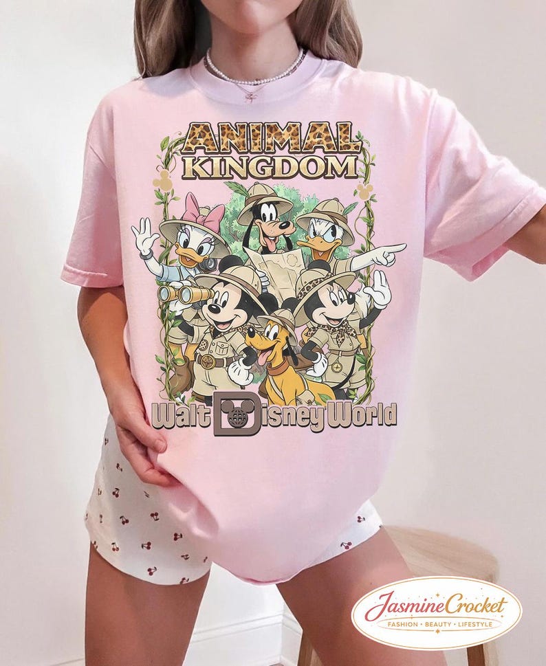 Op de afbeelding: Lichtroze T-shirt met een afbeelding van Disney-figuren in safari-outfits. De tekst "ANIMAL KINGDOM" staat bovenaan, met "Walt Disney World" eronder. Het ontwerp bevat Mickey Mouse, Minnie Mouse, Donald Duck, Daisy Duck, Goofy en Pluto.