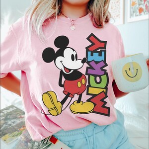May include: Pale pink t-shirt with a Mickey Mouse graphic. The design shows Mickey in his iconic outfit, with the word "Mickey" in vertical, multi-coloured letters. A white mug with a yellow smiley face is also visible.