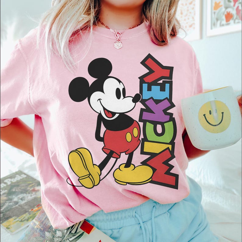 May include: Pale pink t-shirt with a Mickey Mouse graphic. The design shows Mickey in his iconic outfit, with the word "Mickey" in vertical, multi-coloured letters. A white mug with a yellow smiley face is also visible.