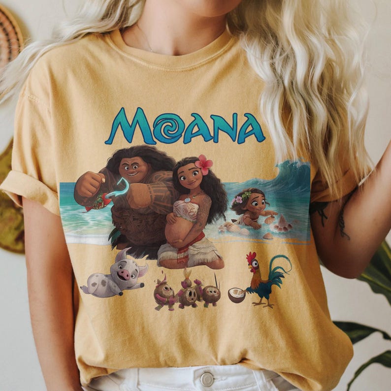 Puede incluir: Camiseta de color amarillo mostaza con la palabra "Moana" en turquesa. La camiseta muestra un gr&aacute;fico colorido de personajes de la pel&iacute;cula animada Moana, incluyendo a Moana, Maui y Hei Hei.