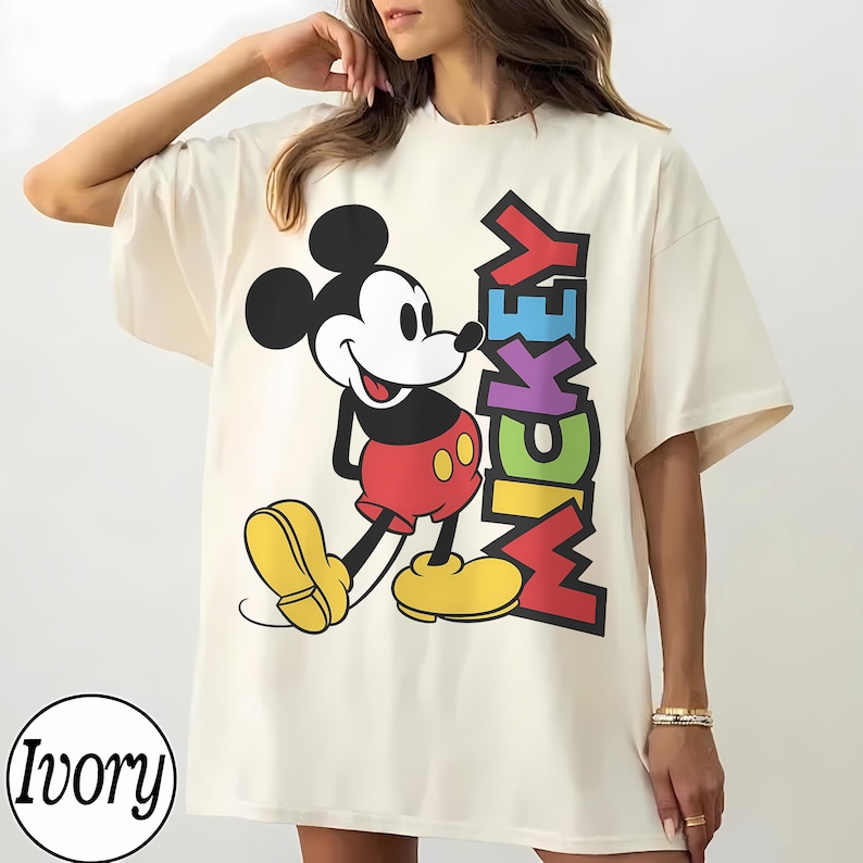 May include: Ivory-coloured t-shirt featuring a large graphic of Mickey Mouse in his classic attire. The word "Mickey" is vertically printed in colourful letters. The shirt has short sleeves and a relaxed fit.