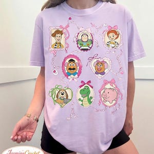 以下が含まれることがあります： ピンクのリボンとリボンで飾られた、額入りの漫画のキャラクターの肖像画が特徴の薄紫色のTシャツ。キャラクターには、ウッディ、バズ・ライトイヤー、ジェシー、ミスター・ポテトヘッドなどが含まれます。シャツの下部には「Jasmine Crocket」の文字があります。