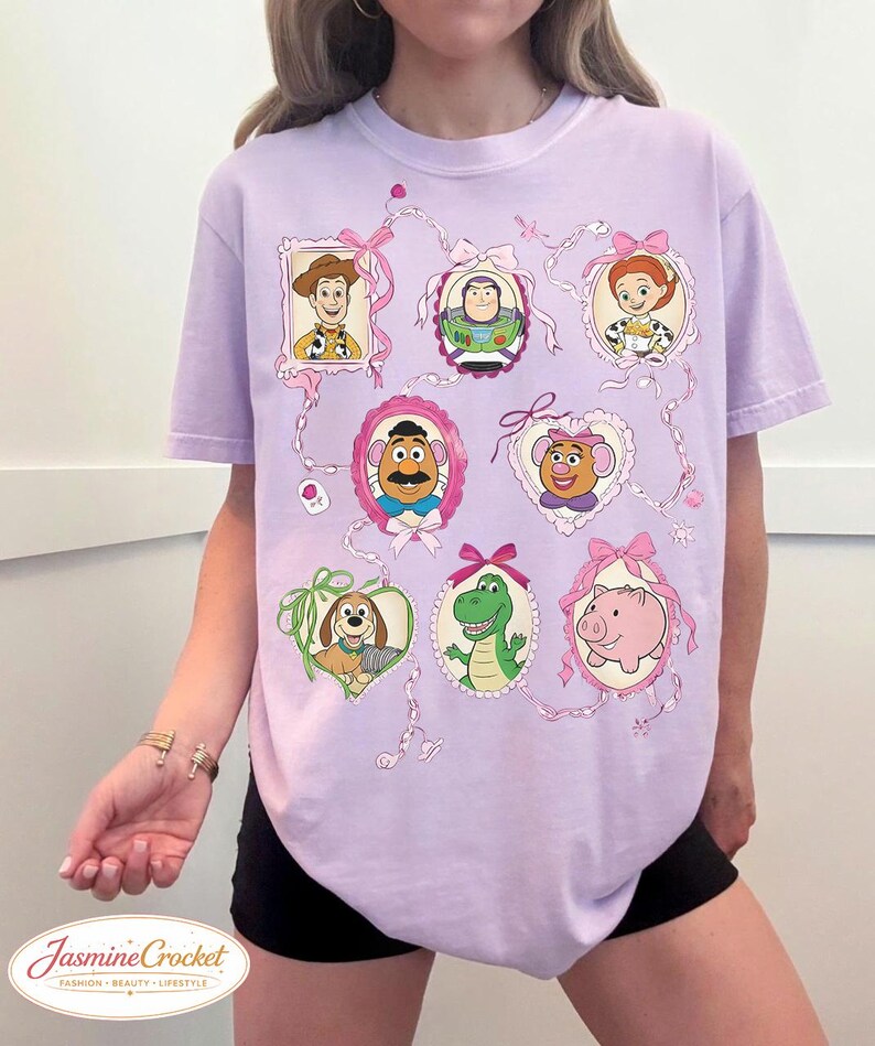 以下が含まれることがあります： ピンクのリボンとリボンで飾られた、額入りの漫画のキャラクターの肖像画が特徴の薄紫色のTシャツ。キャラクターには、ウッディ、バズ・ライトイヤー、ジェシー、ミスター・ポテトヘッドなどが含まれます。シャツの下部には「Jasmine Crocket」の文字があります。