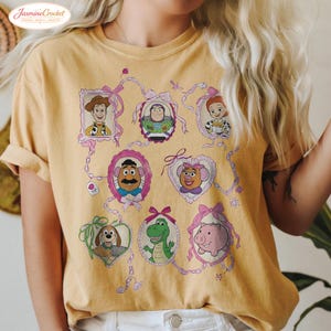 以下が含まれることがあります： マスタードイエローのTシャツ。トイ・ストーリーのキャラクターの額入りポートレートが特徴です。ポートレートはピンクのリボンとリボンでつながっています。キャラクターには、ウッディ、バズ・ライトイヤー、ジェシー、ミスター・ポテトヘッドなどが含まれます。