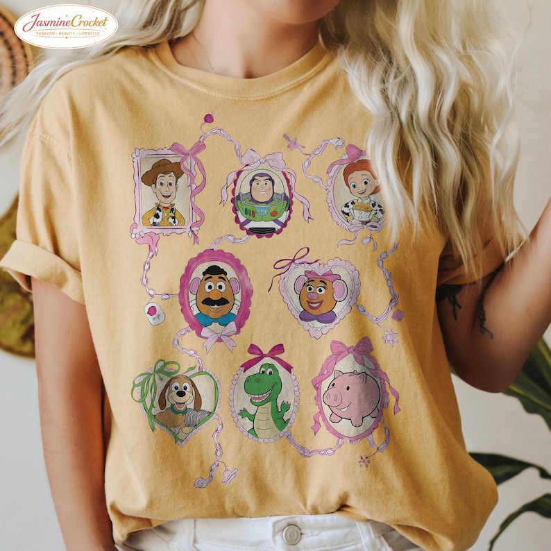 以下が含まれることがあります： マスタードイエローのTシャツ。トイ・ストーリーのキャラクターの額入りポートレートが特徴です。ポートレートはピンクのリボンとリボンでつながっています。キャラクターには、ウッディ、バズ・ライトイヤー、ジェシー、ミスター・ポテトヘッドなどが含まれます。