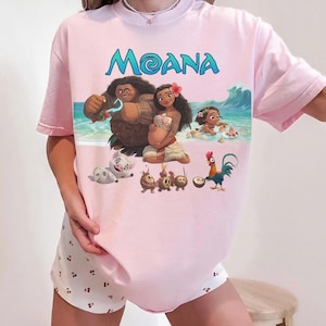 Puede incluir: Camiseta rosa claro con la palabra "MOANA" en letras turquesas. El gr&aacute;fico muestra personajes de la pel&iacute;cula animada, incluyendo a Moana y Maui. La camiseta se combina con pantalones cortos blancos con un peque&ntilde;o estampado de cerezas.
