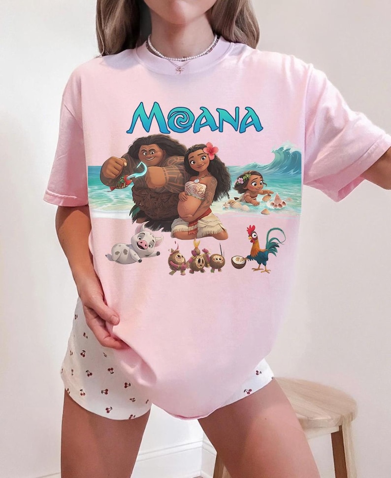 Puede incluir: Camiseta rosa claro con la palabra "MOANA" en letras turquesas. El gr&aacute;fico muestra personajes de la pel&iacute;cula animada, incluyendo a Moana y Maui. La camiseta se combina con pantalones cortos blancos con un peque&ntilde;o estampado de cerezas.