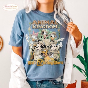 Op de afbeelding: Lichtblauw T-shirt met een afbeelding van Disney-personages in safari-outfits. De tekst "ANIMAL KINGDOM" staat bovenaan, met "Walt Disney World" onderaan. Het ontwerp bevat Mickey Mouse, Minnie Mouse, Donald Duck, Daisy Duck, Goofy en Pluto.
