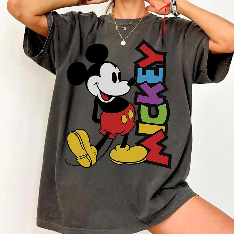 May include: Dark grey t-shirt with a colourful Mickey Mouse graphic and the word "MICKEY" in vertical, multicoloured letters. Mickey is shown in his classic outfit: red shorts, yellow shoes, and black gloves.