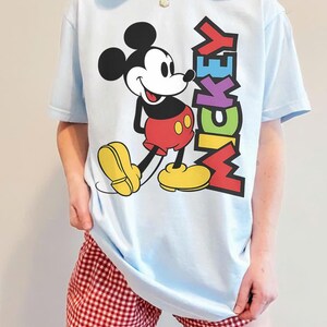 May include: Pale blue t-shirt with a Mickey Mouse graphic. The design shows Mickey in red shorts and yellow shoes, with the word "MICKEY" in various colours. The shirt is worn with red and white checked shorts.