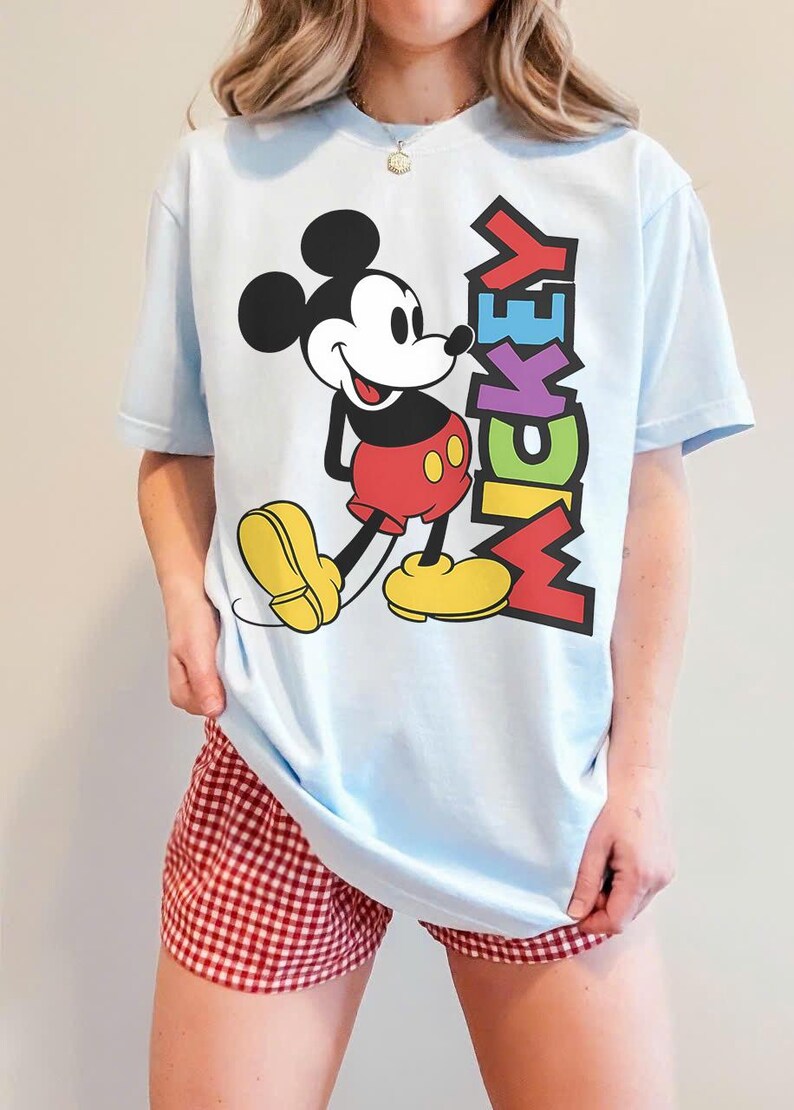May include: Pale blue t-shirt with a Mickey Mouse graphic. The design shows Mickey in red shorts and yellow shoes, with the word "MICKEY" in various colours. The shirt is worn with red and white checked shorts.