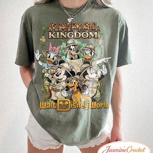 Op de afbeelding: Olijfgroen T-shirt met een afbeelding van Disney-personages in safari-outfits, waaronder Mickey Mouse, Donald Duck en Goofy. De tekst "ANIMAL KINGDOM" en "Walt Disney World" staan op het shirt.