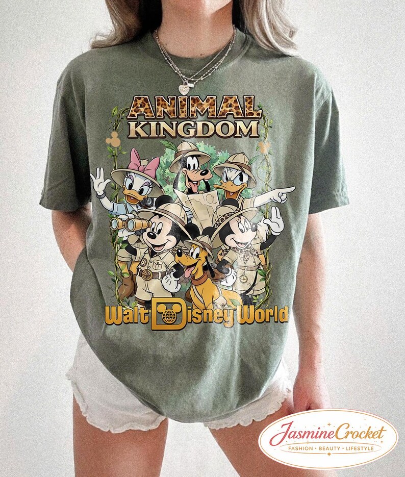Op de afbeelding: Olijfgroen T-shirt met een afbeelding van Disney-personages in safari-outfits, waaronder Mickey Mouse, Donald Duck en Goofy. De tekst "ANIMAL KINGDOM" en "Walt Disney World" staan op het shirt.