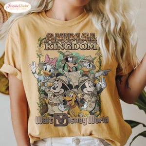 Op de afbeelding: Mosterdgeel T-shirt met een afbeelding van Disney-personages in safari-outfits. De tekst "ANIMAL KINGDOM" staat boven de personages en "Walt Disney World" eronder. Het ontwerp bevat Mickey Mouse, Minnie Mouse, Donald Duck, Daisy Duck, Goofy en Pluto.