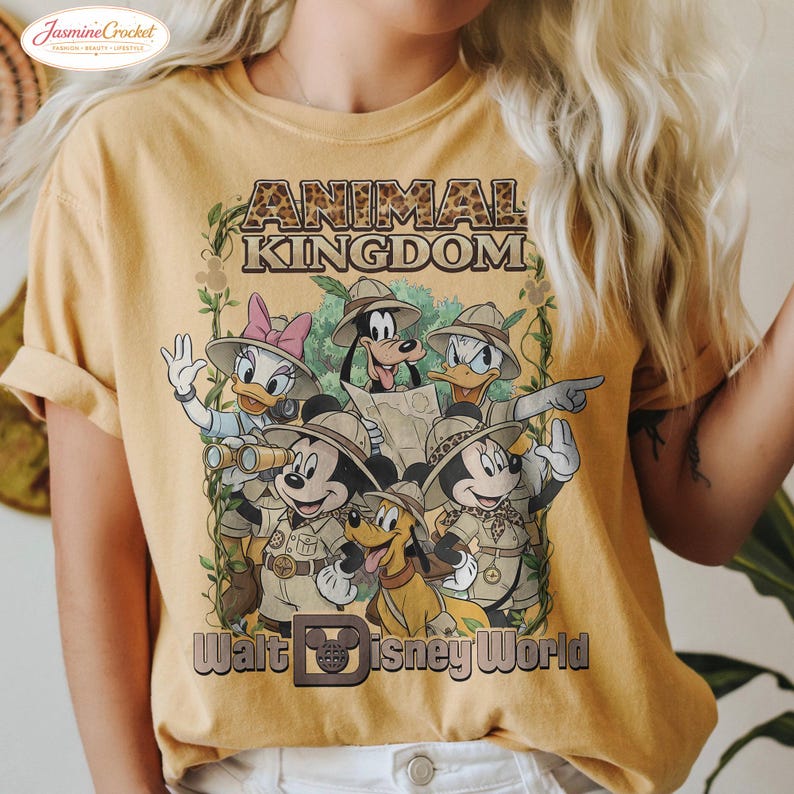 Op de afbeelding: Mosterdgeel T-shirt met een afbeelding van Disney-personages in safari-outfits. De tekst "ANIMAL KINGDOM" staat boven de personages en "Walt Disney World" eronder. Het ontwerp bevat Mickey Mouse, Minnie Mouse, Donald Duck, Daisy Duck, Goofy en Pluto.
