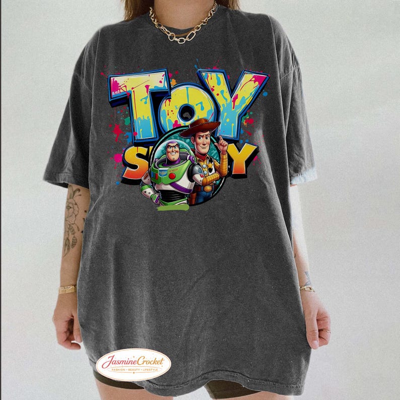 Puede incluir: Camiseta gris oscuro de gran tama&ntilde;o con el texto "TOY STORY" en un dise&ntilde;o colorido estilo grafiti. El gr&aacute;fico incluye im&aacute;genes de Buzz Lightyear y Woody. La camiseta tiene mangas cortas y un corte holgado.