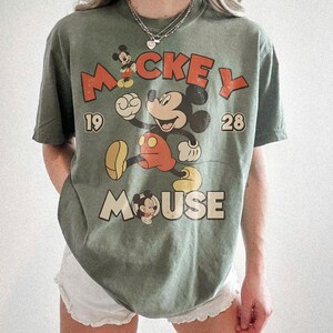 Op de afbeelding: Olijfgroen t-shirt met een vintage Mickey Mouse-ontwerp. Het ontwerp bevat de woorden "Mickey Mouse" en de cijfers "19" en "28". Mickey Mouse wordt afgebeeld in zijn klassieke pose, met een rode broek en gele schoenen.