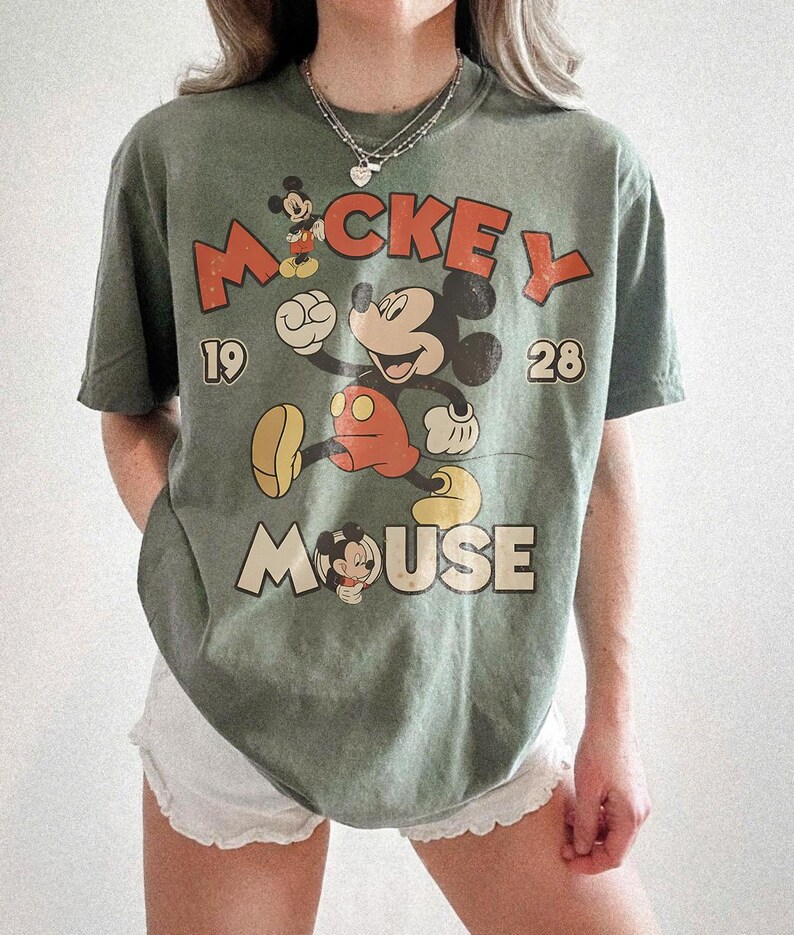 Op de afbeelding: Olijfgroen t-shirt met een vintage Mickey Mouse-ontwerp. Het ontwerp bevat de woorden "Mickey Mouse" en de cijfers "19" en "28". Mickey Mouse wordt afgebeeld in zijn klassieke pose, met een rode broek en gele schoenen.