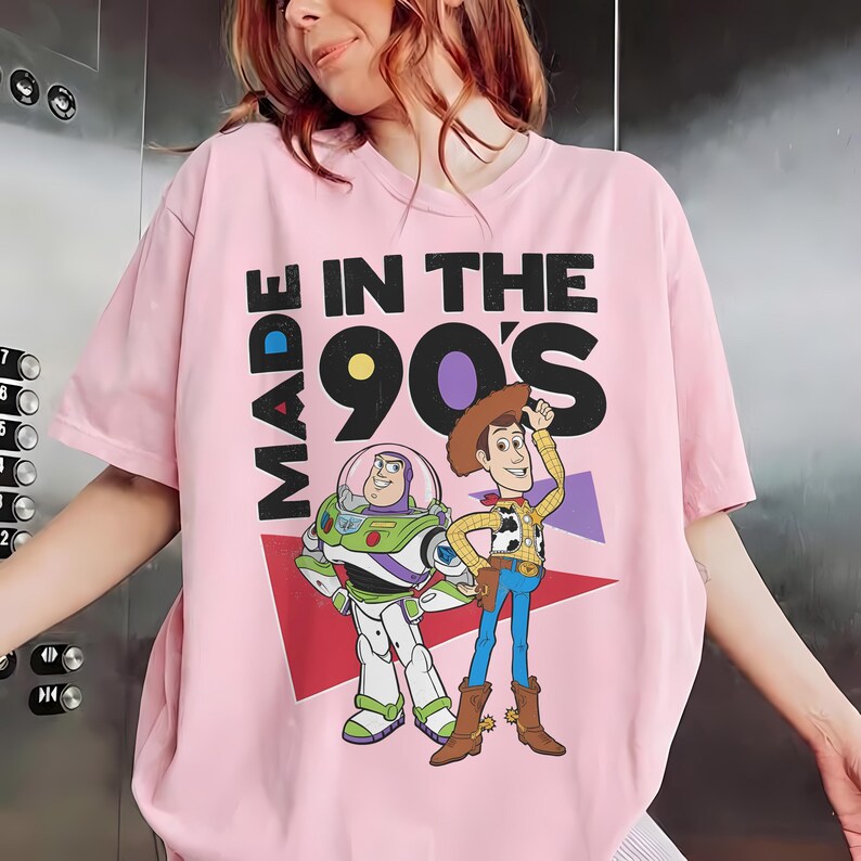 May include: Pale pink t-shirt with the text "MADE IN THE 90'S" and cartoon characters Buzz Lightyear and Woody. The top has a crew neck and short sleeves, suitable for casual wear.