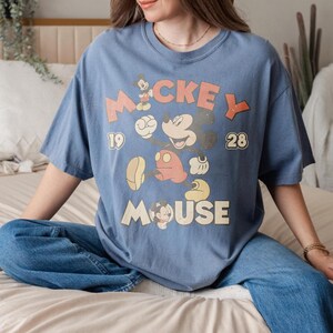 Op de afbeelding: Blauw T-shirt met een vintage Mickey Mouse-ontwerp. Het ontwerp bevat Mickey Mouse, de tekst "Mickey Mouse" en de cijfers "19" en "28". Het shirt heeft een losse pasvorm.