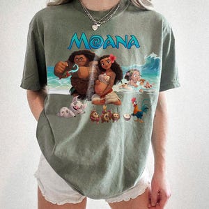 Puede incluir: Camiseta verde oliva con la palabra "Moana" en escritura azul y un gr&aacute;fico colorido. El gr&aacute;fico representa personajes de la pel&iacute;cula animada, incluyendo a Moana, Maui, Pua, Hei Hei y otros personajes en una escena de playa.