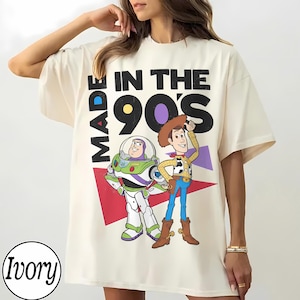 May include: Ivory t-shirt with a graphic of Buzz Lightyear and Woody from Toy Story, and the text "MADE IN THE 90'S". The design includes red and purple geometric shapes.