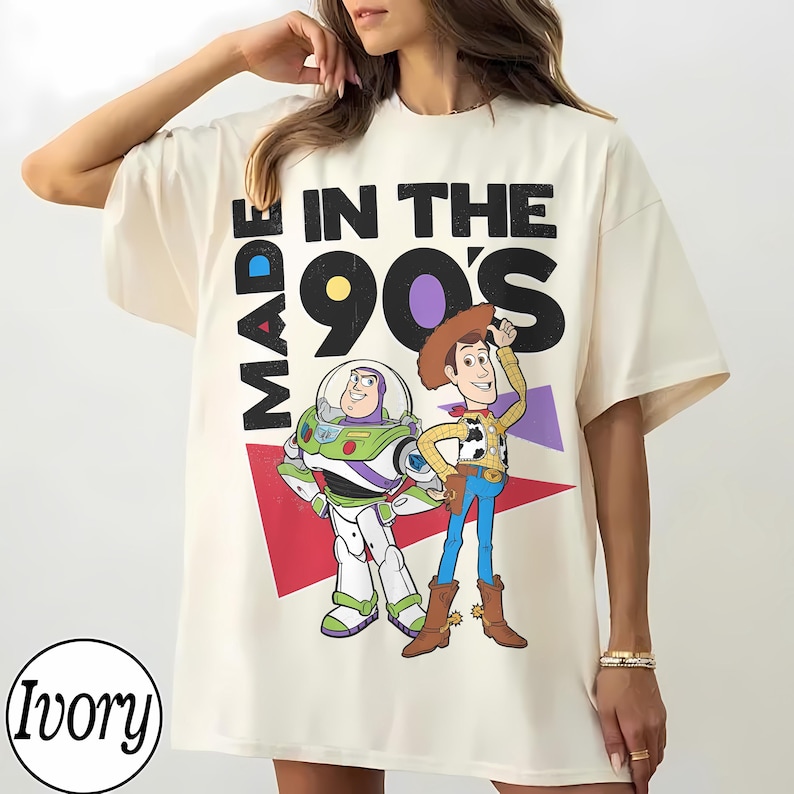 May include: Ivory t-shirt with a graphic of Buzz Lightyear and Woody from Toy Story, and the text "MADE IN THE 90'S". The design includes red and purple geometric shapes.