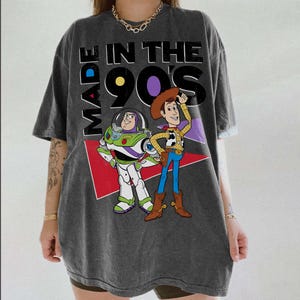 May include: Dark grey t-shirt with the text "MADE IN THE 90'S" and cartoon characters Buzz Lightyear and Woody. The design includes red, purple, and white elements. The shirt has short sleeves and a loose fit.