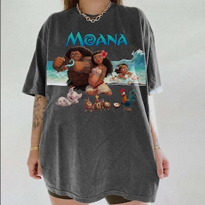 Puede incluir: Camiseta gris oscuro con la palabra "Moana" en turquesa sobre un gr&aacute;fico colorido. El gr&aacute;fico representa personajes de la pel&iacute;cula animada, incluyendo Moana, Maui y Hei Hei, con una ola en el fondo. La camiseta tiene mangas cortas.