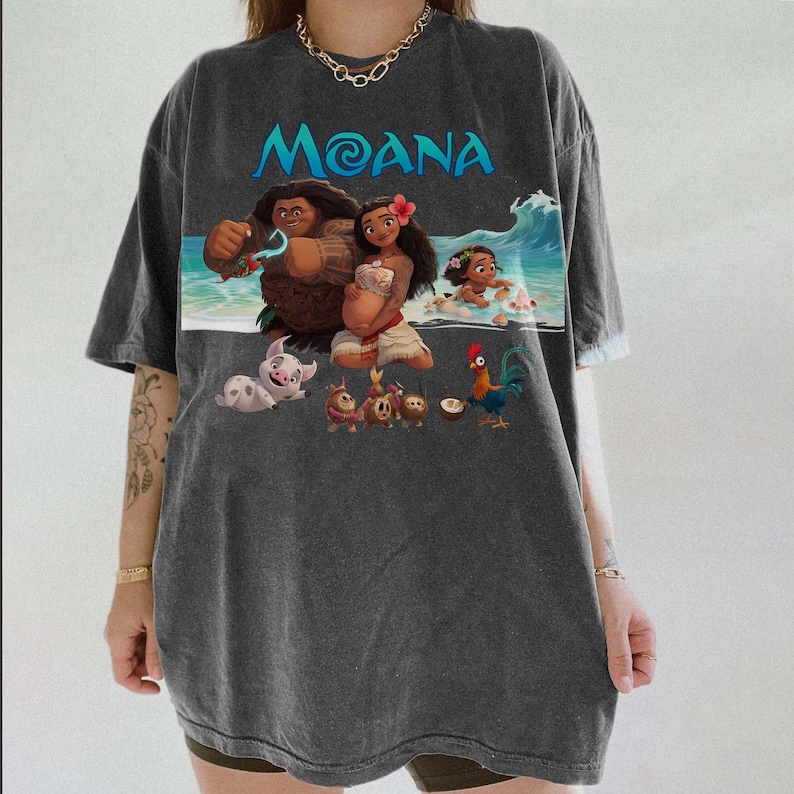 Puede incluir: Camiseta gris oscuro con la palabra "Moana" en turquesa sobre un gr&aacute;fico colorido. El gr&aacute;fico representa personajes de la pel&iacute;cula animada, incluyendo Moana, Maui y Hei Hei, con una ola en el fondo. La camiseta tiene mangas cortas.