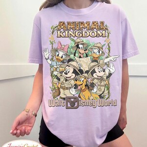 Op de afbeelding: Lila T-shirt met een afbeelding van Disney-personages in safari-outfits, waaronder Mickey Mouse, Donald Duck en Goofy. De tekst "Animal Kingdom" en "Walt Disney World" staan boven en onder de personages.