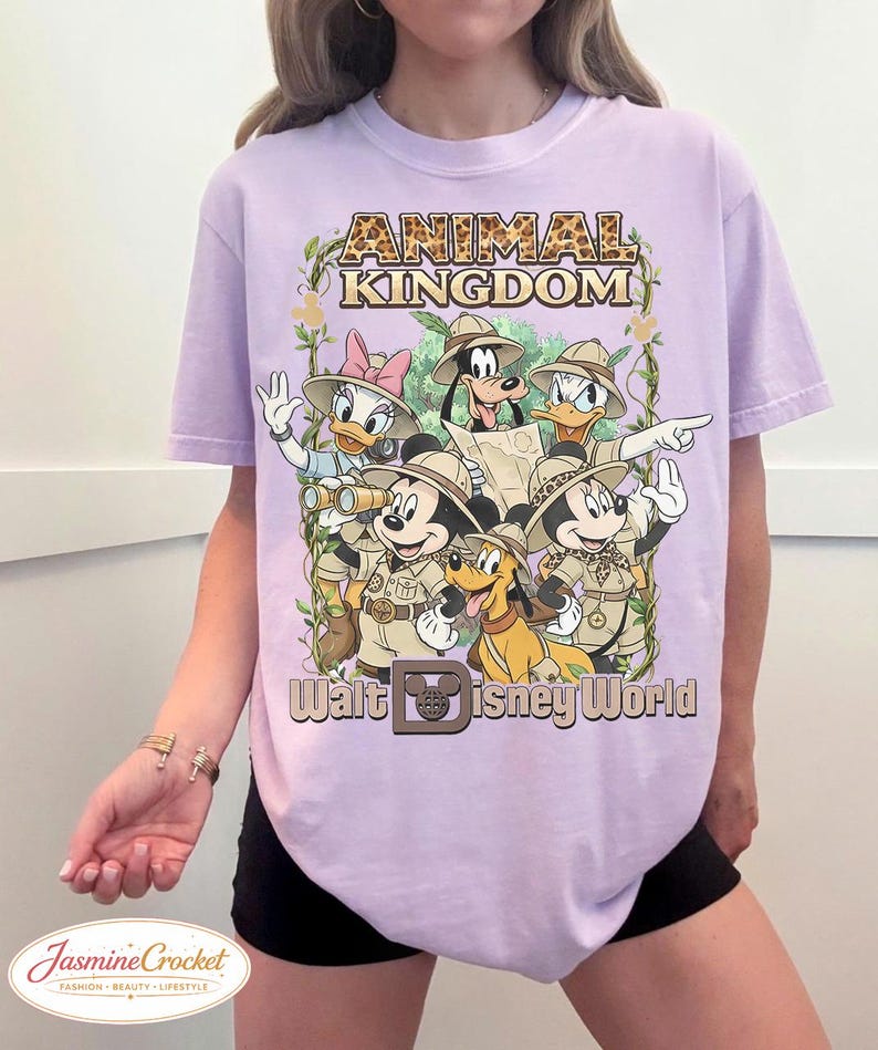 Op de afbeelding: Lila T-shirt met een afbeelding van Disney-personages in safari-outfits, waaronder Mickey Mouse, Donald Duck en Goofy. De tekst "Animal Kingdom" en "Walt Disney World" staan boven en onder de personages.